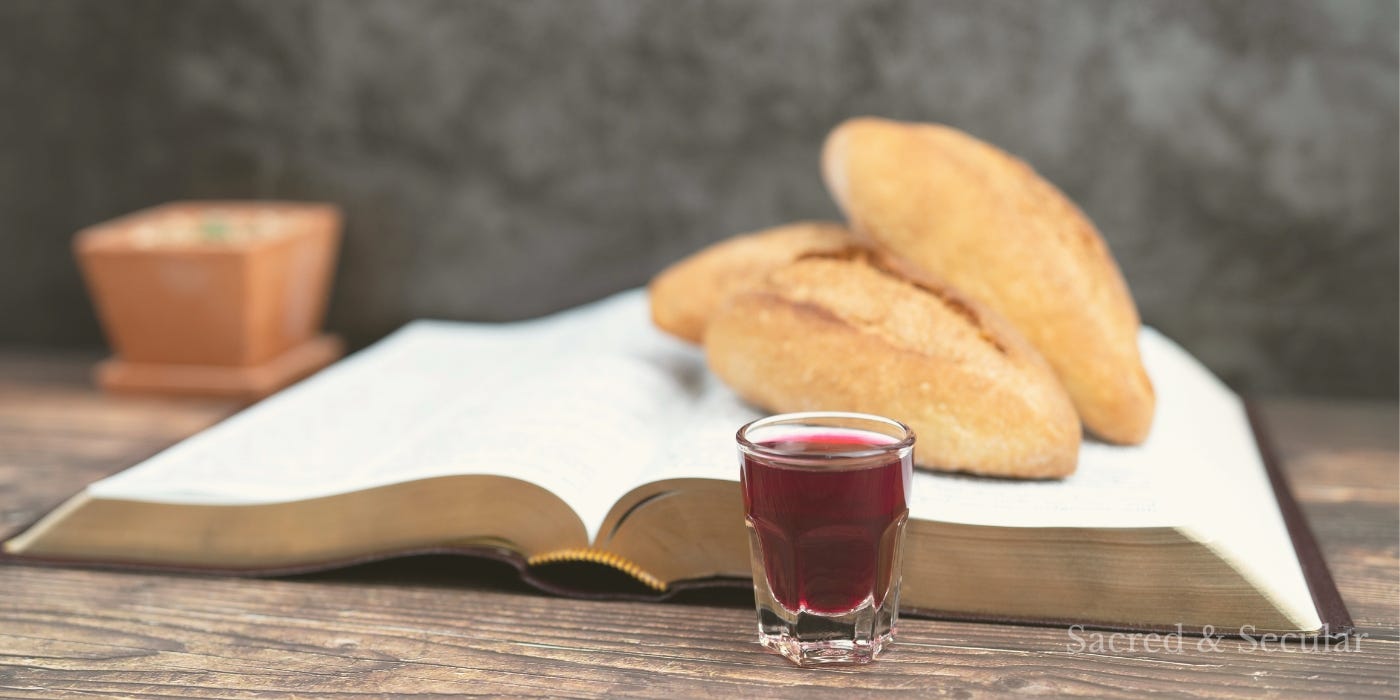 A simple wooden table with an open Bible, pieces of bread resting on its pages, and a small glass of red wine in the foreground, softly lit in a calm, reflective setting.