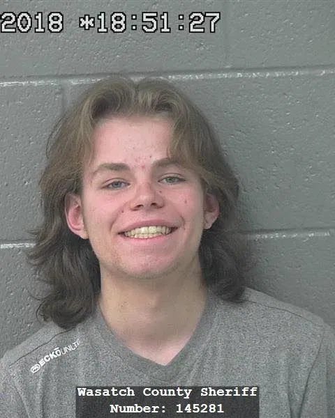 An adult smiling in a gray shirt, against a gray wall, with a computer time stamp at the top of the image, "2018 *18:51:27" and, at the bottom of the image, "Wasatch County Sheriff Number: 145281." An adult smiling in a gray shirt, against a gray wall, with a computer time stamp at the top of the image, "2018 *18:51:27" and, at the bottom of the image, "Wasatch County Sheriff Number: 145281."