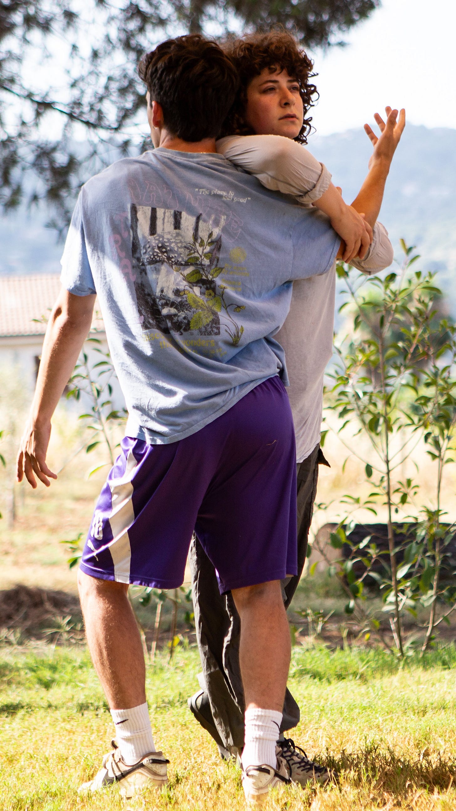 On a brightly lit hillside a muscular dancer wearing a lilac T-shirt, purple shorts and trainers is clasped by a curly haired dancer in a pink sweater who can only partially be seen.