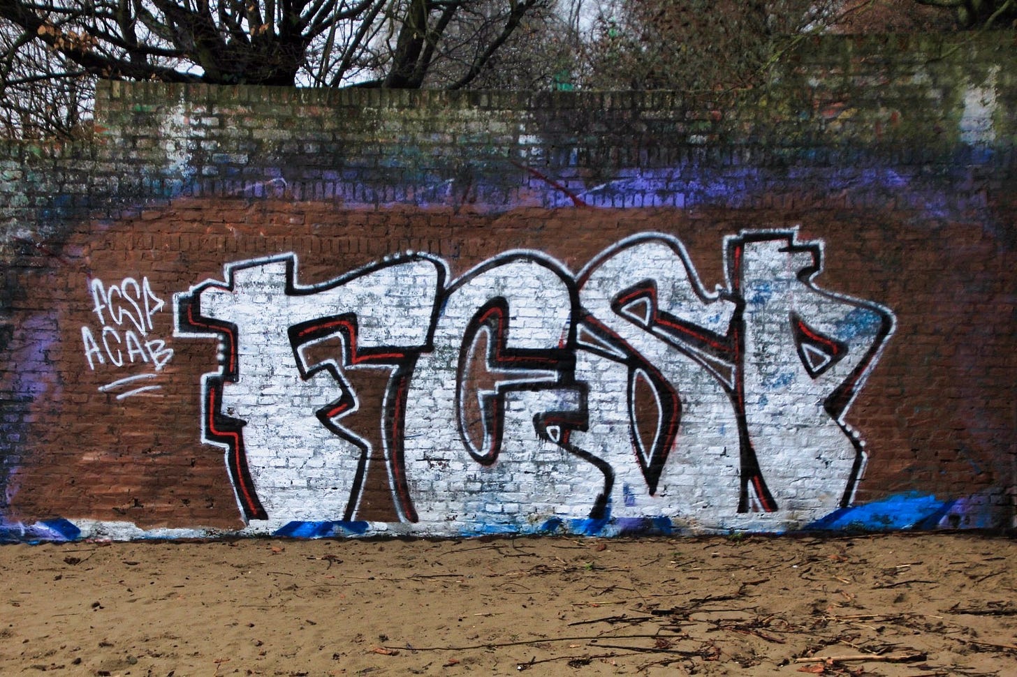Graffiti auf einer alten Backsteinmauer im Freien. In der Bildmitte dominieren große, wild geschwungene Wildstyle-Buchstaben in Weiß mit schwarzen Konturen und roten Akzenten — lesbar als „FCSP" (vermutlich FC St. Pauli). Links daneben steht in kleiner, weißer Schrift ebenfalls „FCSP" und darunter „ACAB". Der Hintergrund der Mauer ist rotbraun, im oberen Bereich mit einem violett-blauen Farbstreifen. Am Boden verläuft ein blauer Farbrand. Hinter der Mauer ragen kahle Bäume in einen bedeckten Winterhimmel. Der Boden vor der Mauer ist sandig und mit trockenen Ästen bedeckt.