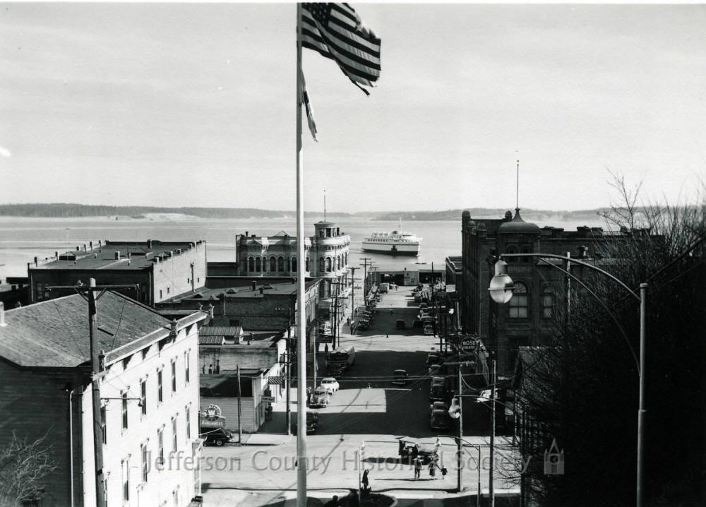 Black-and-white historic photograph looking downhill over downtown Port Townsend toward the waterfront and Port Townsend Bay. A tall flagpole with an American flag stands prominently in the foreground above the Haller Fountain area.