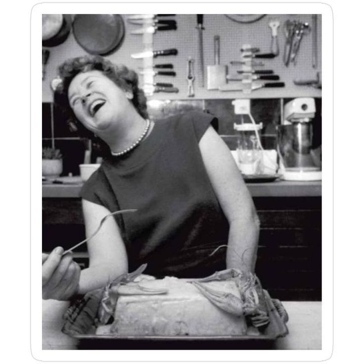 Black-and-white photo of Julia Child laughing in her kitchen while preparing a roast, surrounded by vintage cookware and utensils.