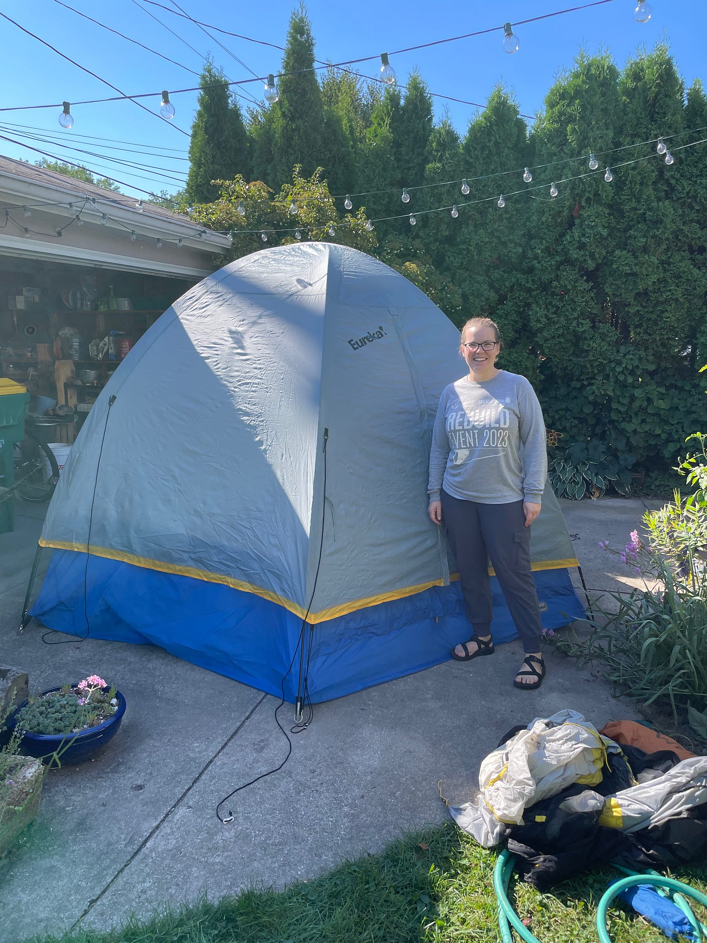 The author is posed next to a grey tent that is taller than she is, with an open garage and a hedge of trees in the background.