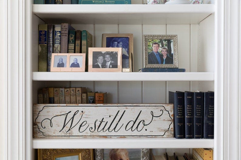 May include: A white bookshelf with three shelves. The top shelf has books and a framed photo of a couple. The middle shelf has books and a wooden sign that says 'We still do.' The bottom shelf has books and two framed photos.