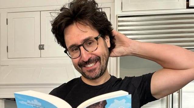 Adam Ragusea smiles for the camera while holding a book. He is a late-30s or early-40s year old male with dark hair, dark beard scruff, and glasses.