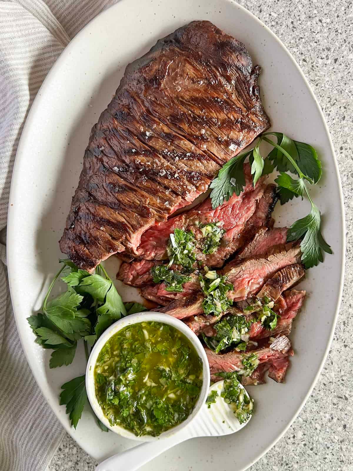 Whole bavette steak with slices cut on a white platter with a small bowl of chimichurri sauce.
