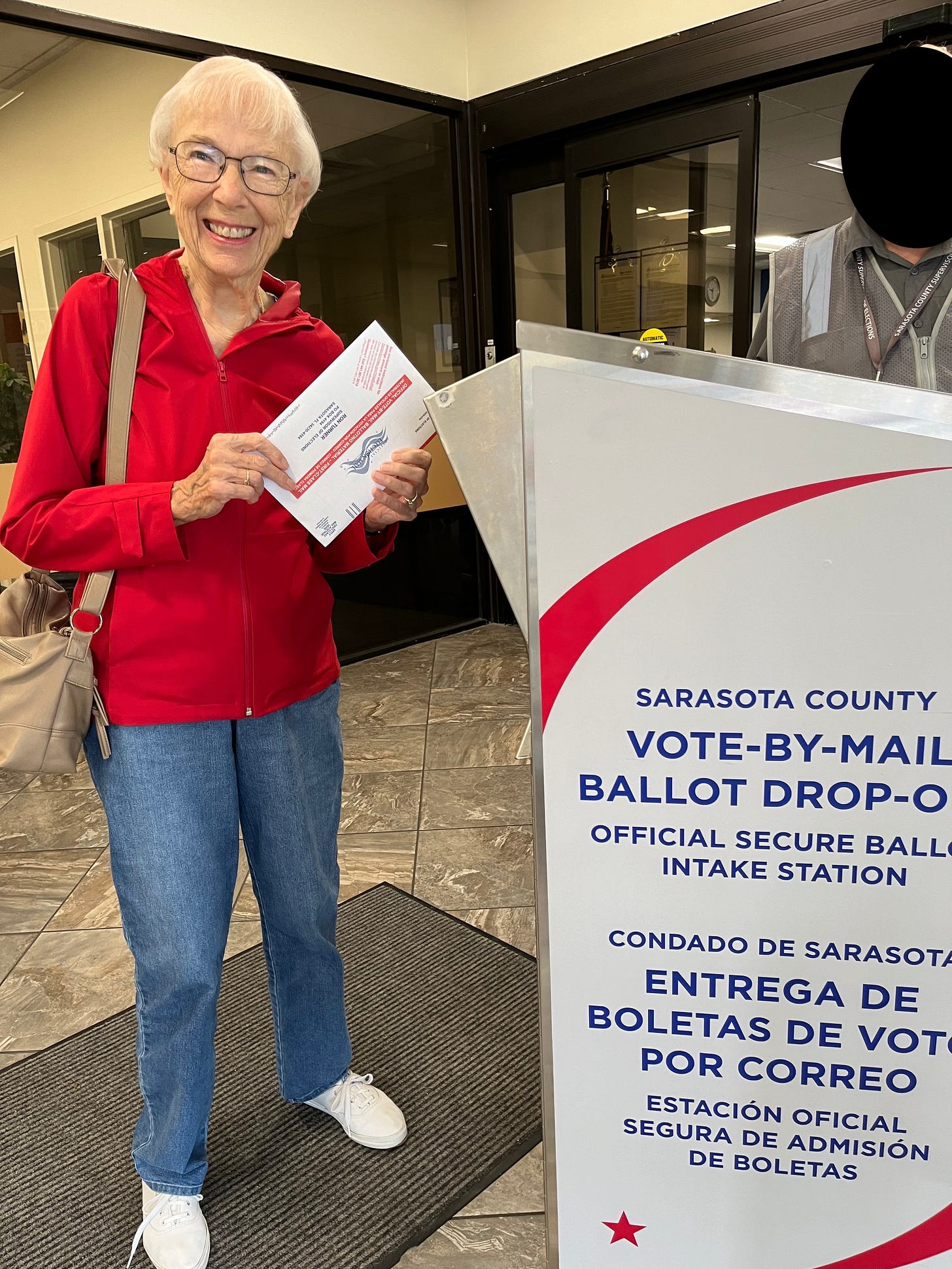 A very pretty elderly woman wearing glasses and smiling widely holds the vote-by-mail ballot she is about to put into a box that reads on the side "Sarasota County Vote-By-Mail ballot drop-off Official secure ballot intake station," with the message repeated in Spanish below. She is dressed in a red raincoat, blue mom jeans, and very white Keds sneakers. Her handbag is over one shoulder.