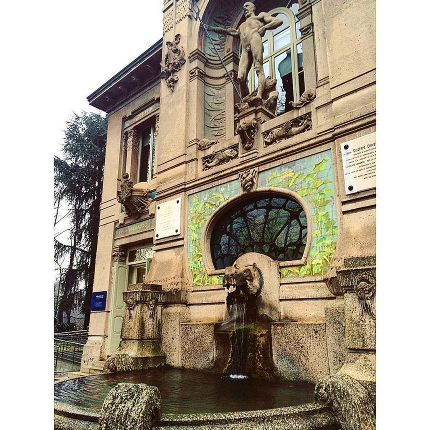 a fountain in front of a building with statues on the side and windows above it
