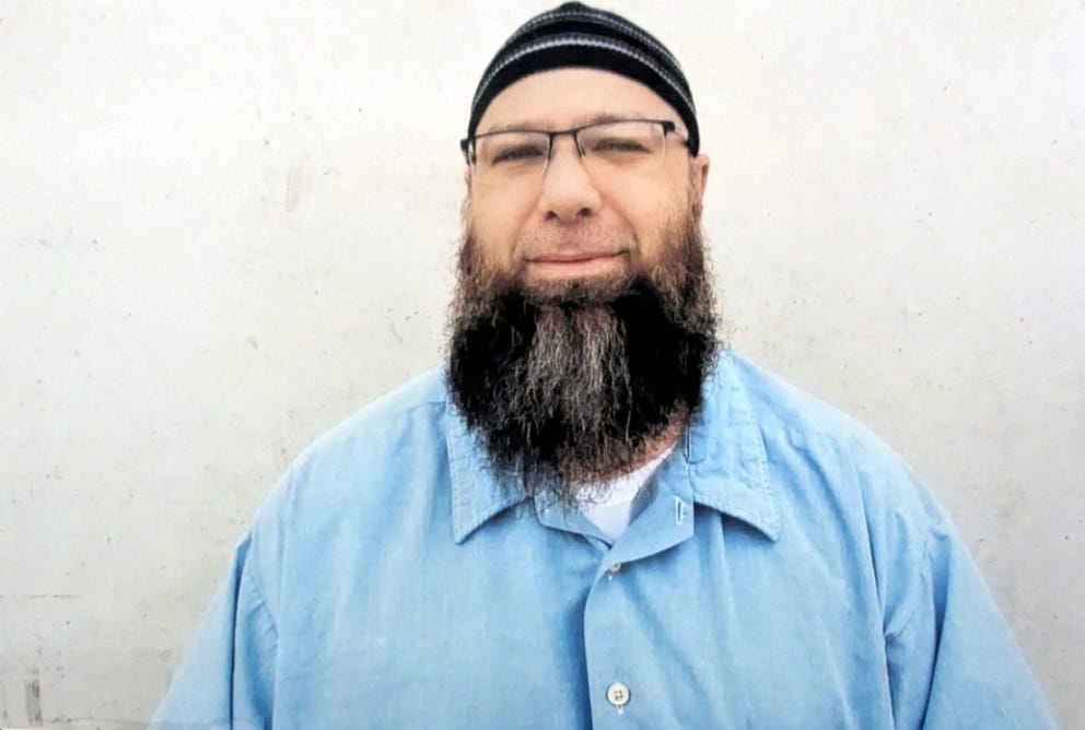 Talib Muwahhid looks into the camera, wearing a hat, glasses, a beard and a blue buttondown shirt.