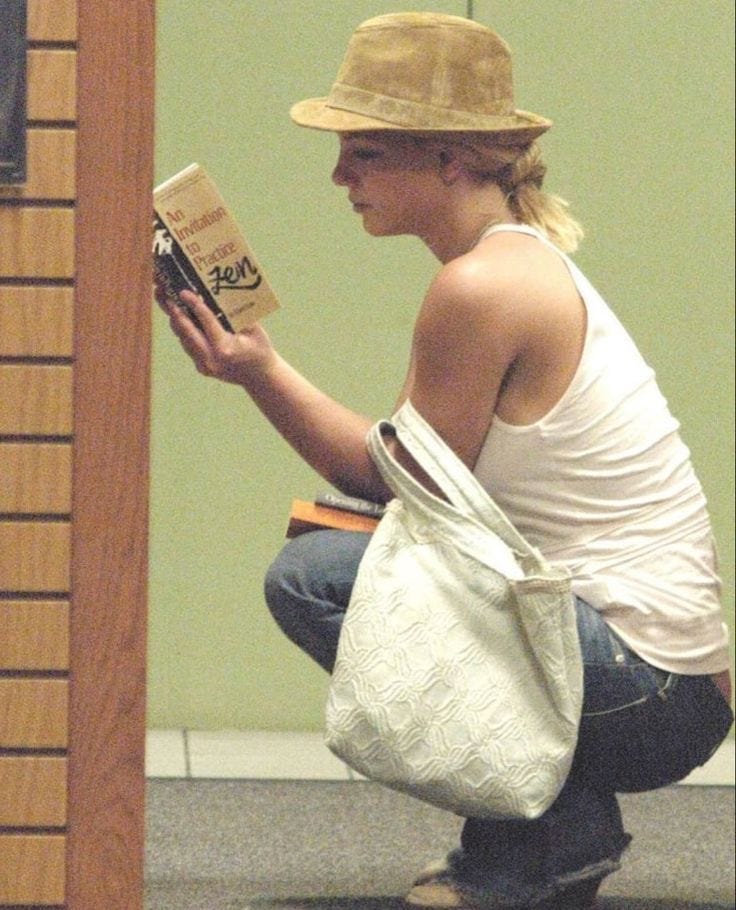 This may contain: a woman kneeling down reading a book while holding a white bag and wearing a hat
