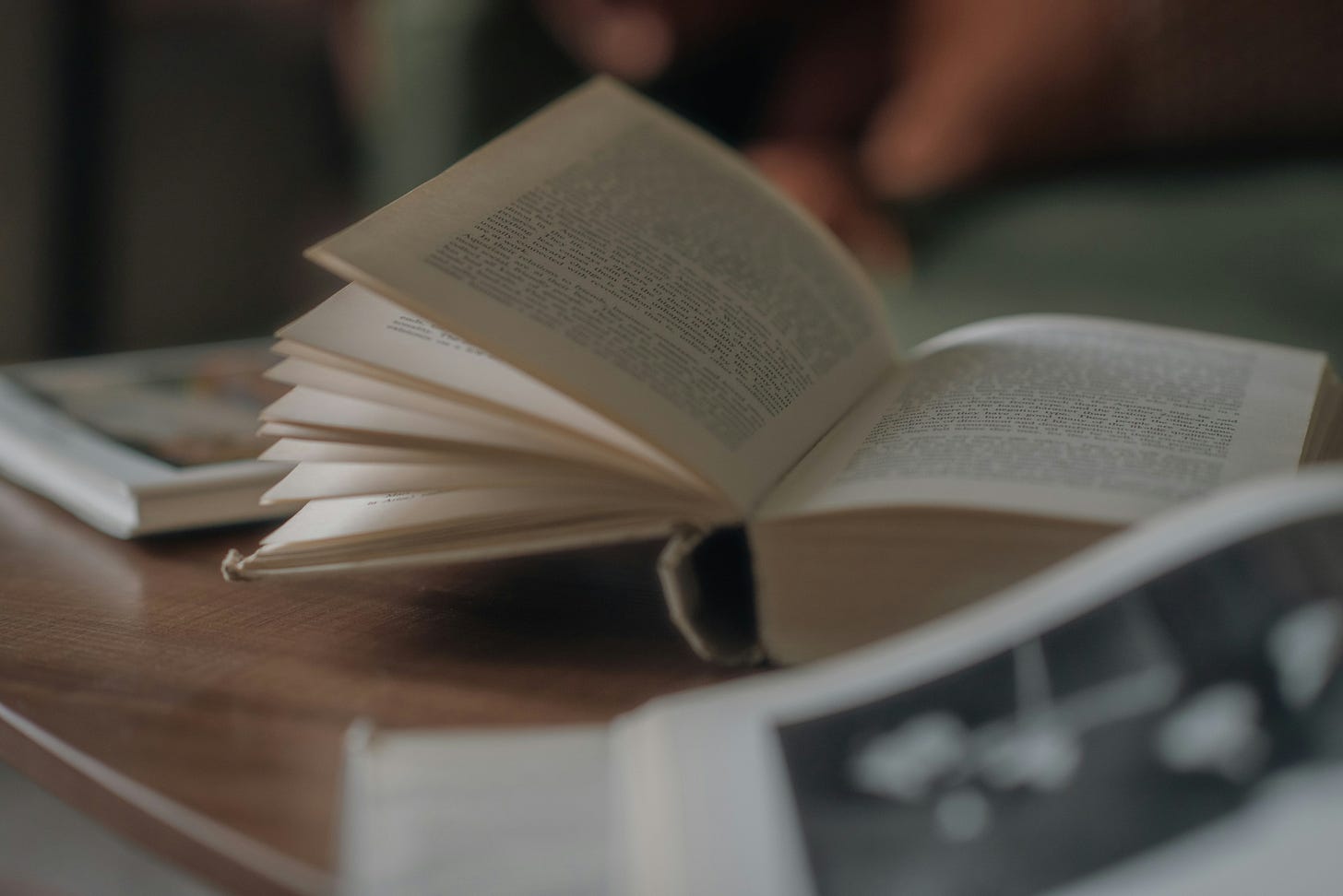 An open book with a white cover on a brown wooden table. There is another book behind it and in front of it but is blurred out.