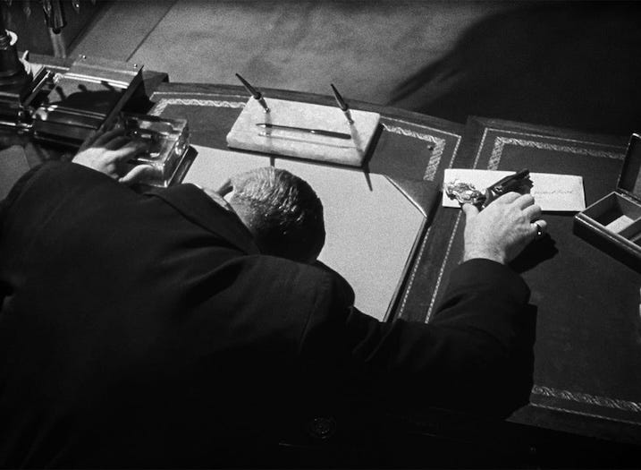 A man lies motionless over an ornate desk, one hand clutching a pistol, the other near a glass inkwell. A stark overhead light casts deep shadows, emphasizing the film’s fatalism and mood.