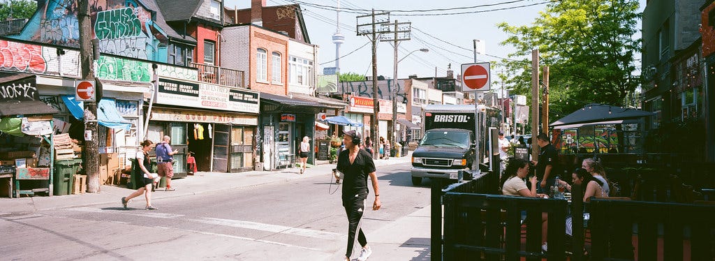 Kensington Market. a street with market stands, people walking around, some sitting outside a cafe. the buildings are small and funky and there is graffiti on the roofs. Kensington Market. a street with market stands, people walking around, some sitting outside a cafe. the buildings are small and funky and there is graffiti on the roofs.