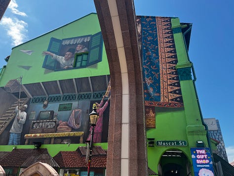 Top left photo is a green street sign that reads Haji Lane. Middle top photo is a white and blue shopping bag with Loka Made in print letters. Top right photo is a crowded street with shops on both sides of the lane. Bottom left photo is a street mural on Muscat Street. Bottom middle photo is of the Sultan Mosque. Bottom right photo is a gray mailbox and a stack of postcards being fed into the open slot. 