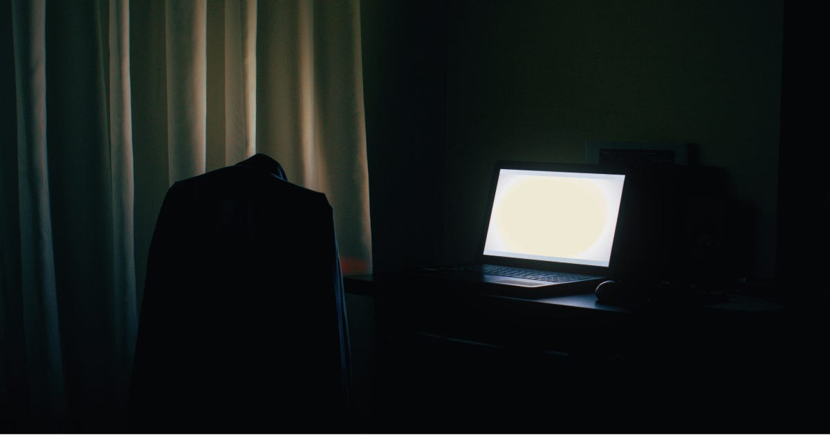A dark room lit by an open laptop beside an empty, draped chair.