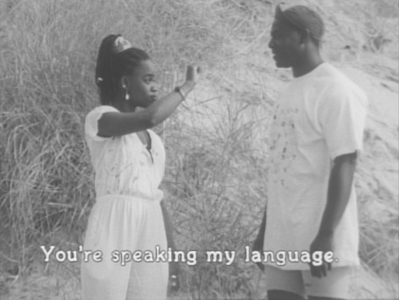 Standing on a beach, a Black woman holds up her hand, signing to a Black man. A caption at the bottom of the screen reads: “You’re speaking my language.”