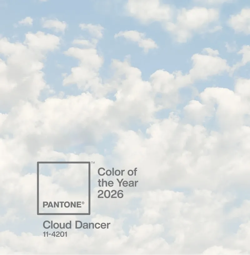 Promotional image from Pantone showing a large Pantone color chip graphic labeled “Cloud Dancer 11-4201” framing a portion of a cloudy sky. The chip’s rectangular outline isolates a soft, pale gray-blue area of clouds, visually equating the atmospheric color with its printed code. The caption identifies it as Pantone’s Color of the Year 2026 and frames the image as a sky translated into a standardized, reproducible color system.