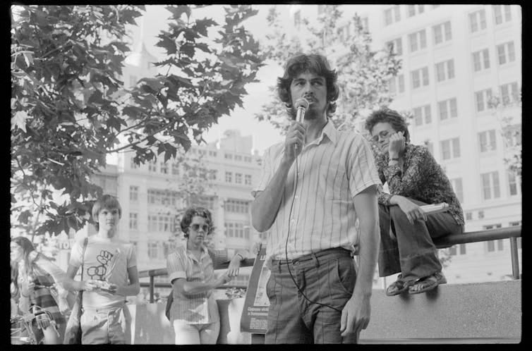 a man talking into a microphone, in a shirt and trousers, with onlookers listening a man talking into a microphone, in a shirt and trousers, with onlookers listening