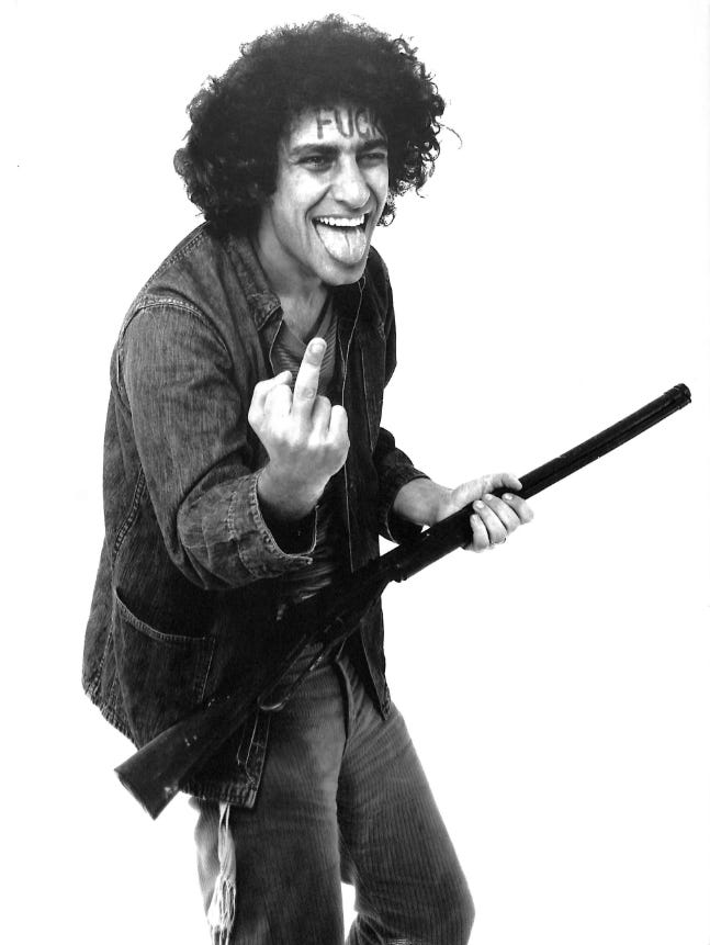 A man in double denim and a T shirt smiles with his tongue out, poses with a toy rifle and flips the bird. He has the word FUCK painted on his forehead