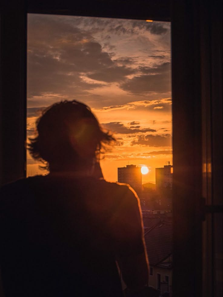 This may contain: a person standing in front of a window with the sun setting behind them and buildings