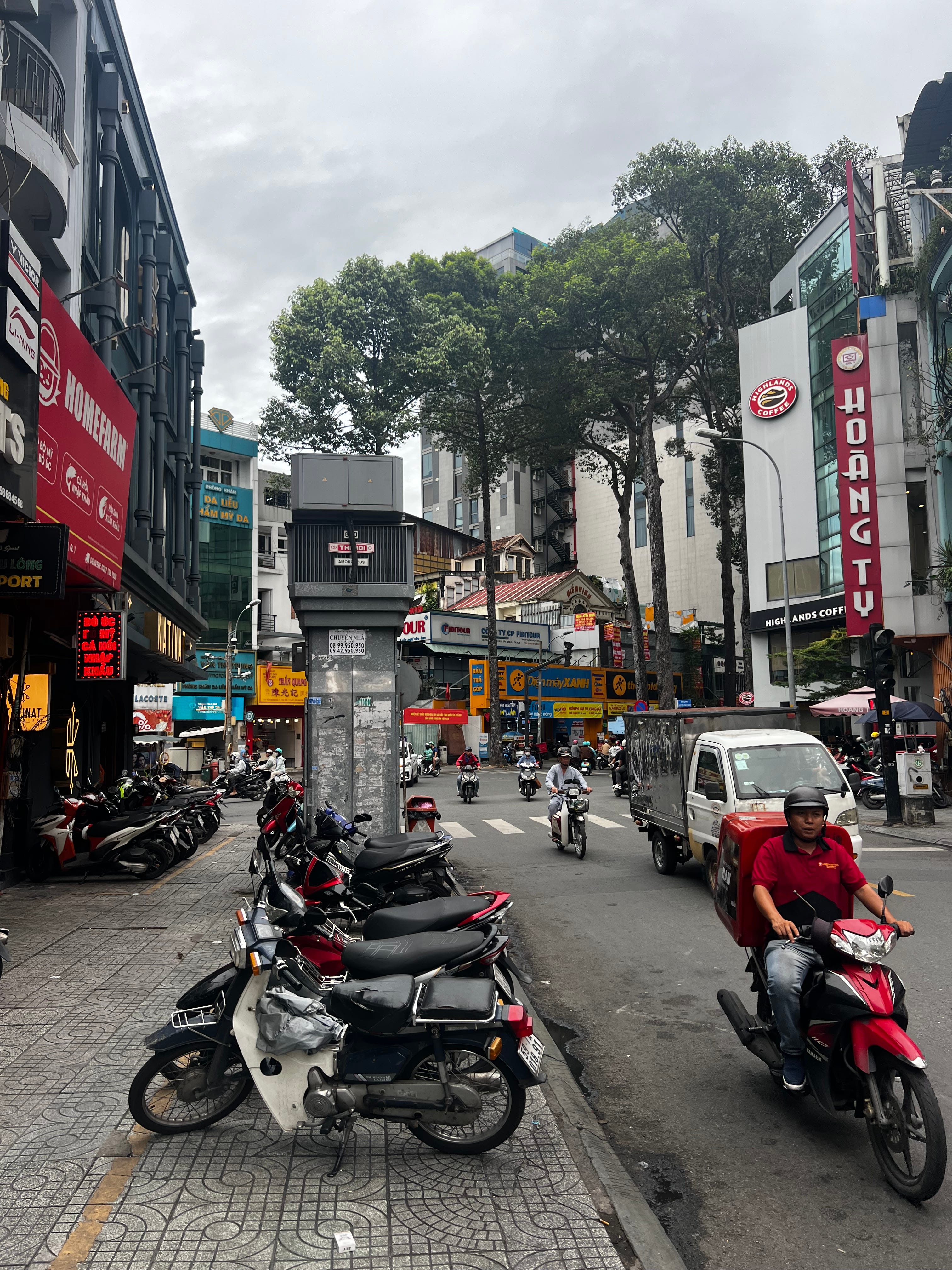 Street scene in Ho Chi Minh City showing motorcycles and scooters navigating a busy intersection under an overcast sky. Tall trees line the street, and colorful storefront signs in Vietnamese and English advertise various businesses including Highlands Coffee. A white and red scooter is parked in the foreground, while a delivery driver on a red motorbike passes through the crosswalk. Multi-story buildings with mixed commercial and residential use line both sides of the street.