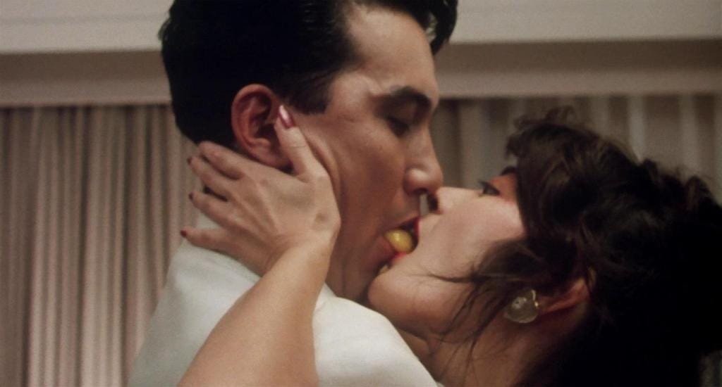 A couple intimately kiss with eyes closed, sharing an egg yolk between their lips. The scene is passionate, set against soft, neutral-coloured curtains.