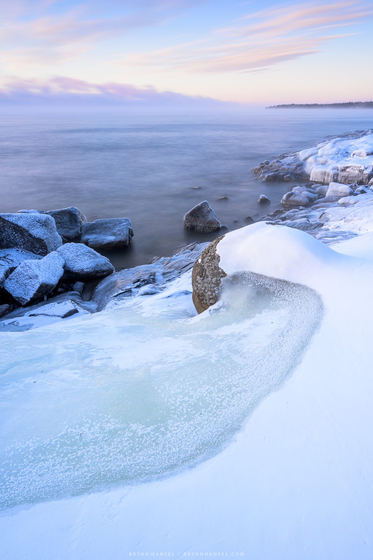 Ice and snow mix on a curved line and point toward muted pink clouds over Lake Superior. 