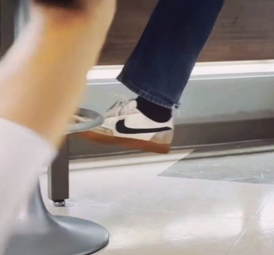 Still from Challengers with Patrick's leg extended, wearing jeans and black socks and Nike Killshots, about to scoot Art's stool before he sits down
