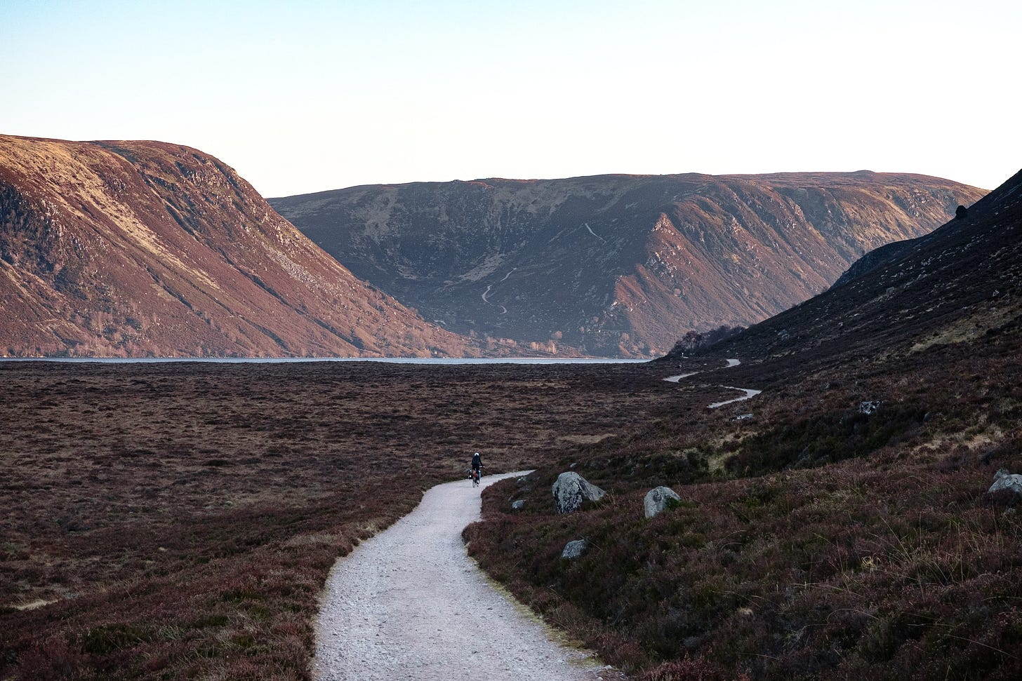 A distant cyclist rides along a narrow path toward Loch Muick, surrounded by steep valley slopes. The barren hills are dusted with heather, giving the scene an expansive, scale-less feel reminiscent of Icelandic wilderness.