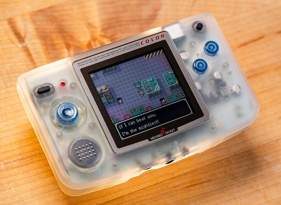 A NeoGeo Pocket Color console on a wooden surface displays a game scene with text: "If I can beat you, I'm the mightiest!"