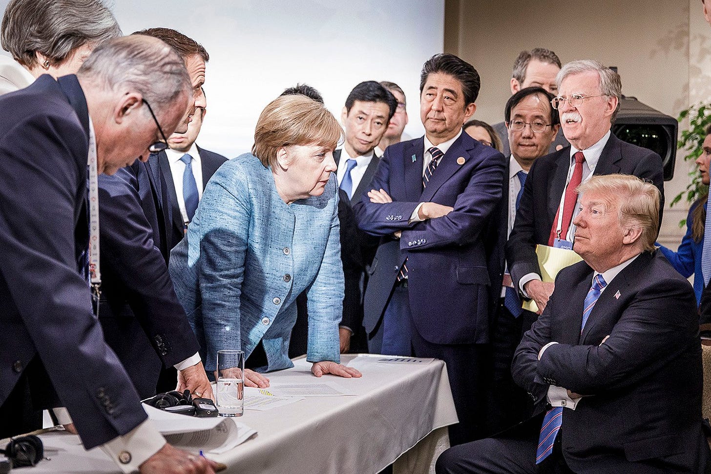 Merkel leans over a table with her arms braced on its surfce to deliberate with U.S. president Donald Trump, who is seated facing her with his arms crossed, surrounded by world leaders. They are surrounded by other world leaders in suits, mostly men, some talking and some listening.