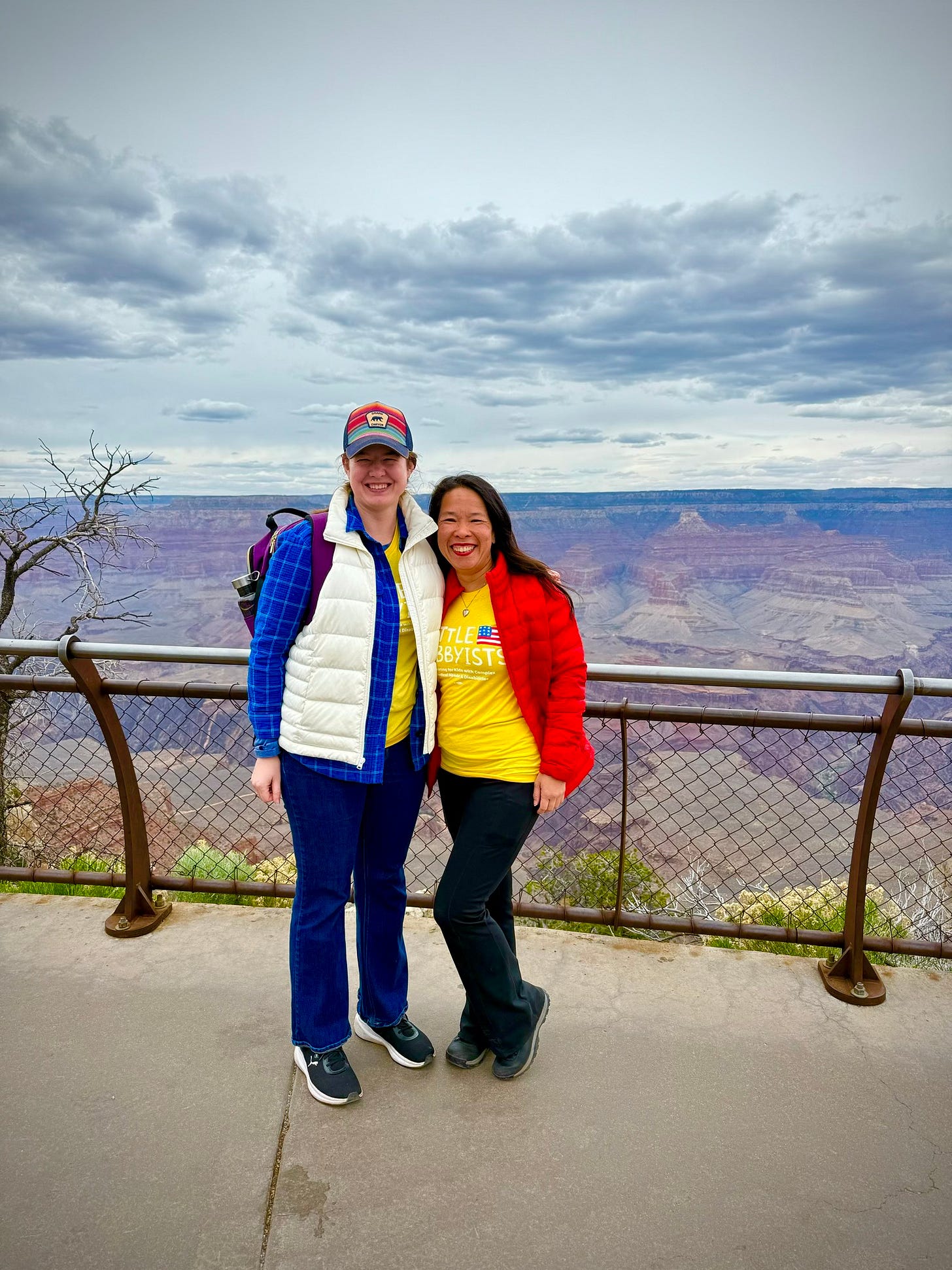 A Caucasian woman blue flannel and white vest and a Chinese woman in a red jacket and yellow shirt stand in front of a metal railing. Behind them is the vista of red rock layers of the Grand Canyon.