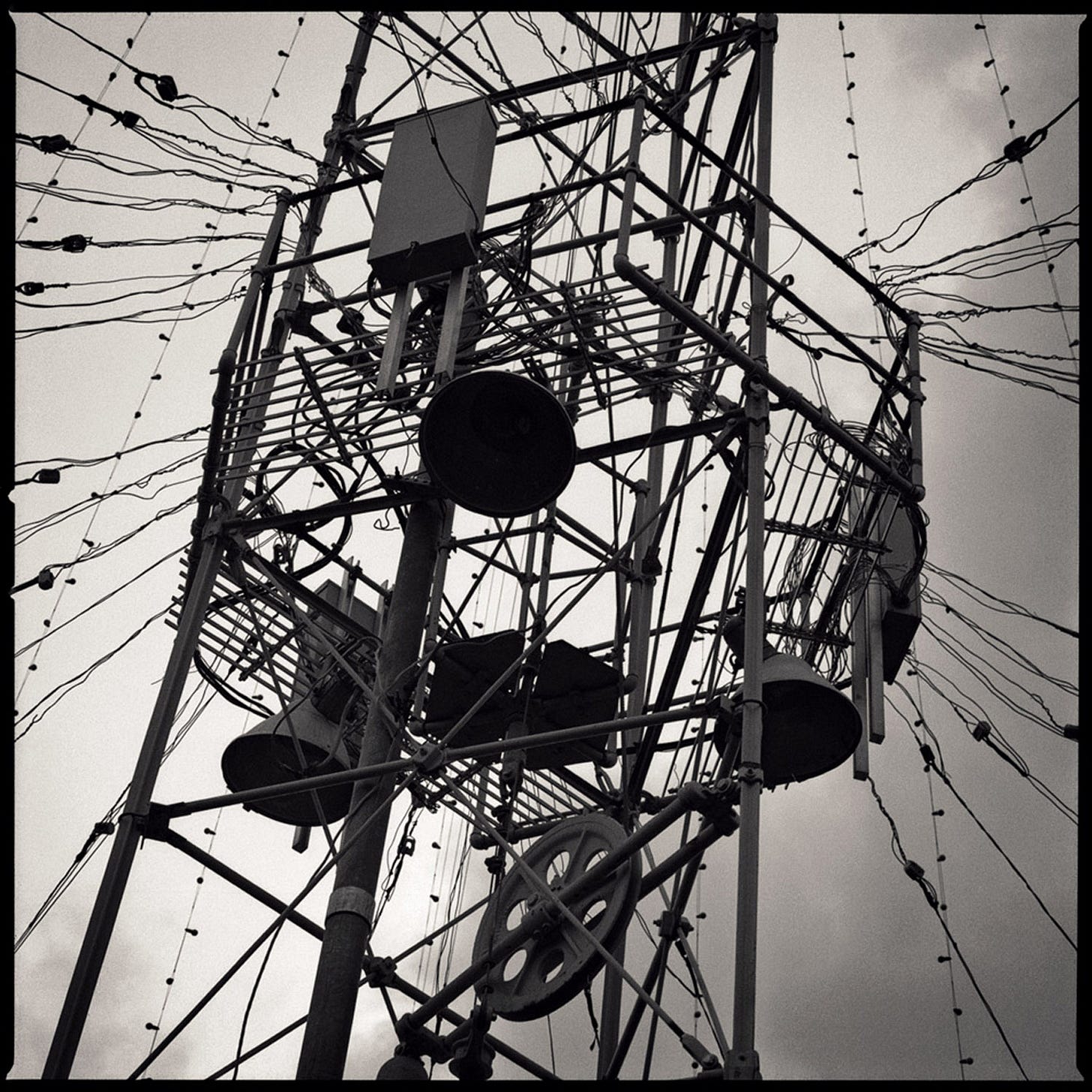 One of Austin's nineteenth-century moonlight towers photographed from below against a clouded sky, horn loudspeakers and rigging mounted in the steel lattice, strings of lamps radiating outward — the tower that anchors the Zilker Holiday Tree. Austin, Texas. Black and white. Photograph by Sean Perry from the series Transitory.