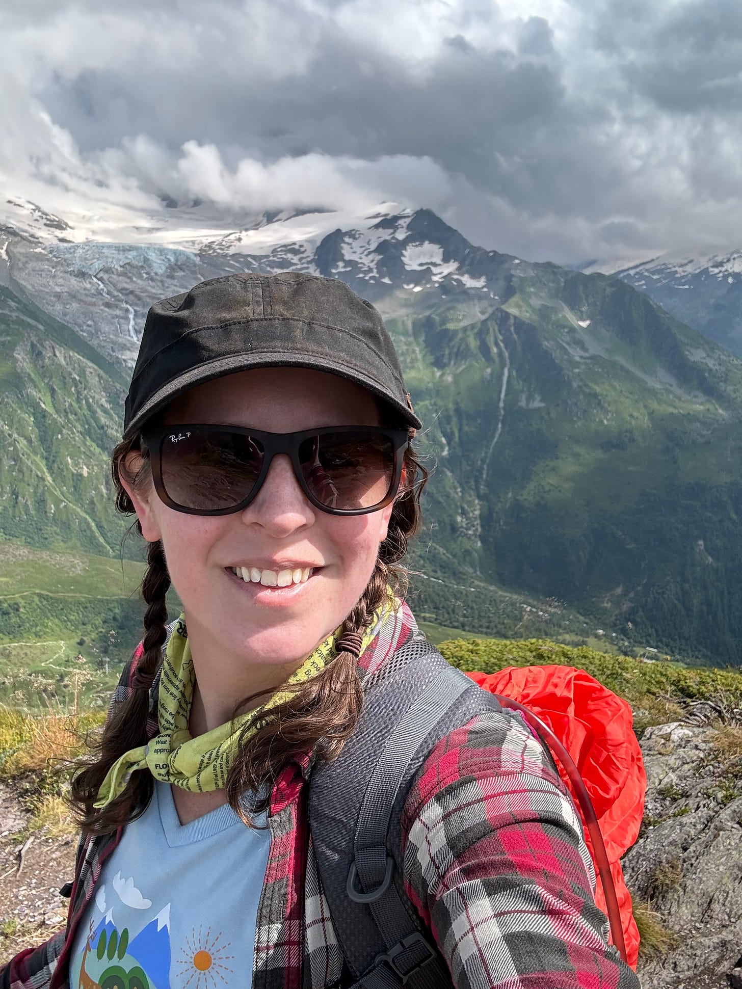 A white woman with brunette piggy tails (also she's an adult) is smiling at the camera with a snowcapped mountain in the background. She's probably on day 9 of a 10 day hike at this point and very sweaty and tired but her hat and sunglasses make it hard to tell.