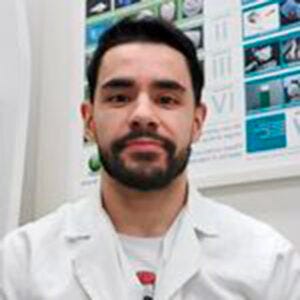 A imagem contém um homem, ele tem cabelo e barba escuros e usa jaleco