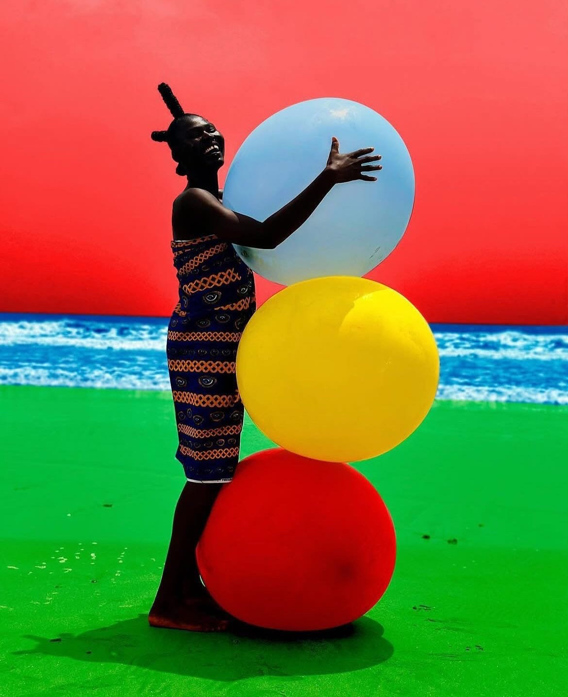 A highly saturated, high-contrast photograph of a smiling Black person with short, sculptural braided hair, wrapped in a dark patterned fabric. They are standing on a solid green floor against a stark, solid bright red background, joyfully wrapping their arms around a vertical stack of three oversized spheres. The spheres are colored light blue (top), bright yellow (middle), and deep red (bottom).