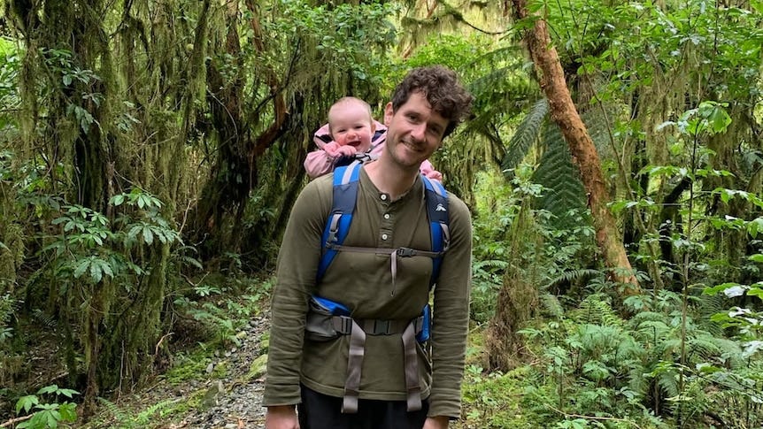 Robbie Arnott smiling on a lush walking trail with his infant daughter strapped in a carrier.