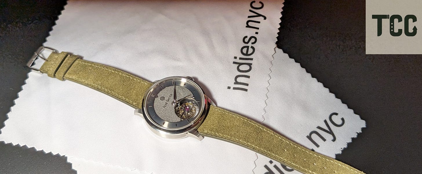 Close-up of a Rogé Bastide wristwatch with visible tourbillon complication and a suede green strap, placed atop a white microfiber cloth printed with the “indies.nyc” logo.
