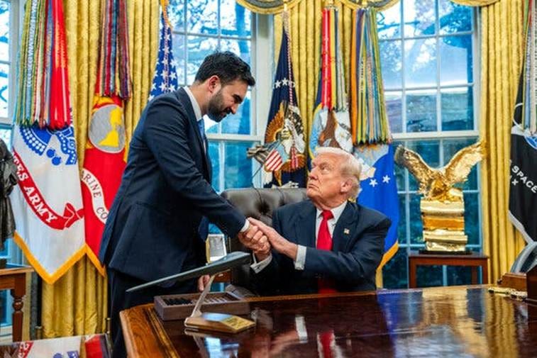 Zohran Mamdani stands above a seated Donald Trump as they shake hands in front of an array of flags in the Oval Office.