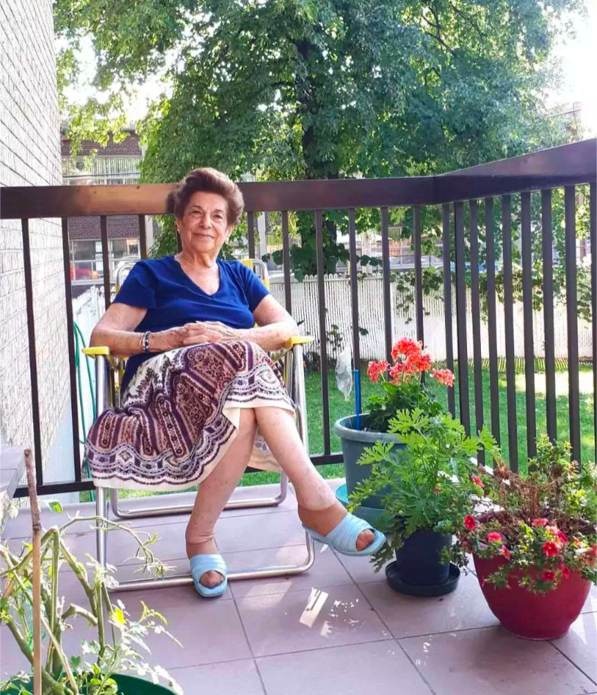 My grandmother sitting in a folding chair on her balcony surrounded by flower pots. She is wearing a royal blue tshirt, a patterned skirt and blue slippers. Her smile suggests she is proud and content.