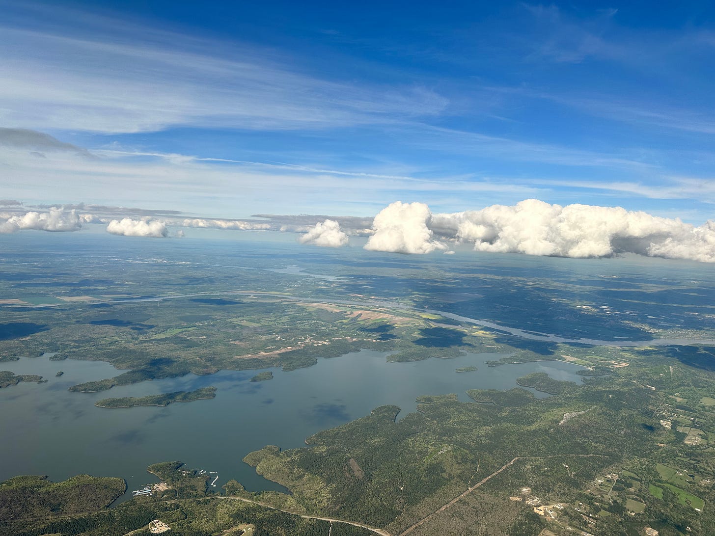 View from airplane overlooking Little Rock Arkansas region at cloud level. Water and green below, blue sky above, flat-bottomed white clouds splitting the view horizontally in the middle. 