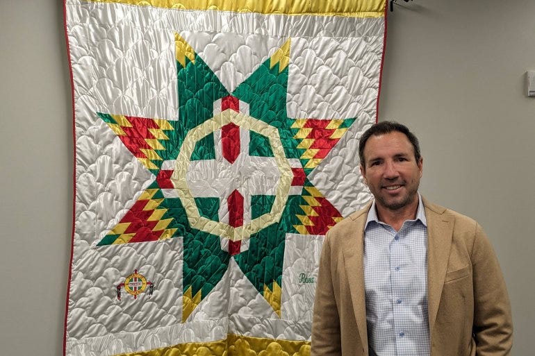 A man in a tan suit jacket smiles at the camera while standing in front of a quilted piece of white, yellow, green, and red fabric mounted to the wall behind him. A man in a tan suit jacket smiles at the camera while standing in front of a quilted piece of white, yellow, green, and red fabric mounted to the wall behind him.