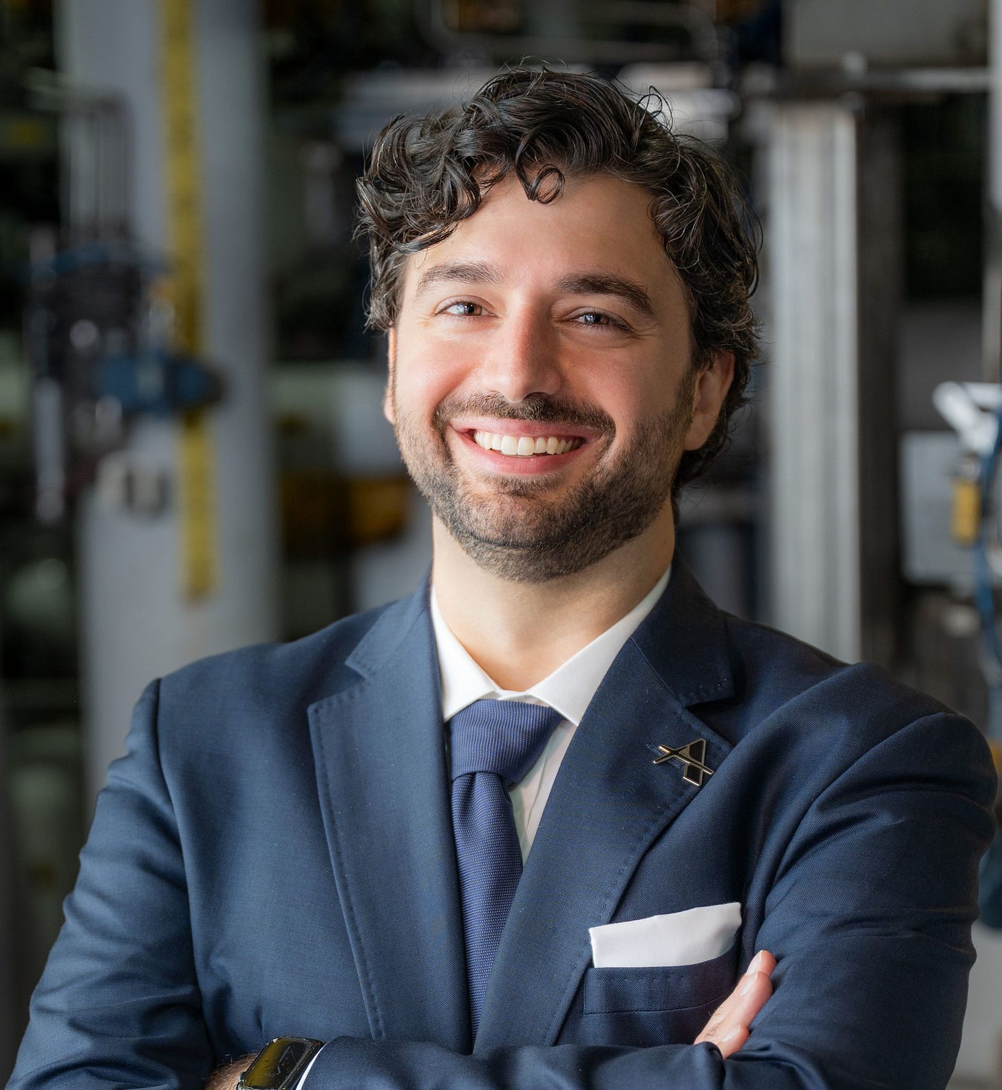 Paul Keutelian, CTO and Co-Founder of Applied Atomics, in a navy suit with an Applied Atomics lapel pin, arms crossed, standing in front of piping and equipment at the IST facility.