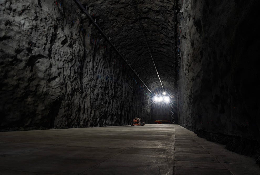 Túnel subterrâneo de grande profundidade com paredes de rocha escura e irregular, iluminado por potente luz branca no topo central, com piso de concreto cinzento e um pequeno veículo de mineração amarelo e laranja estacionado à distância, criando uma composição simétrica que evidencia a escala monumental da caverna. Túnel subterrâneo de grande profundidade com paredes de rocha escura e irregular, iluminado por potente luz branca no topo central, com piso de concreto cinzento e um pequeno veículo de mineração amarelo e laranja estacionado à distância, criando uma composição simétrica que evidencia a escala monumental da caverna.