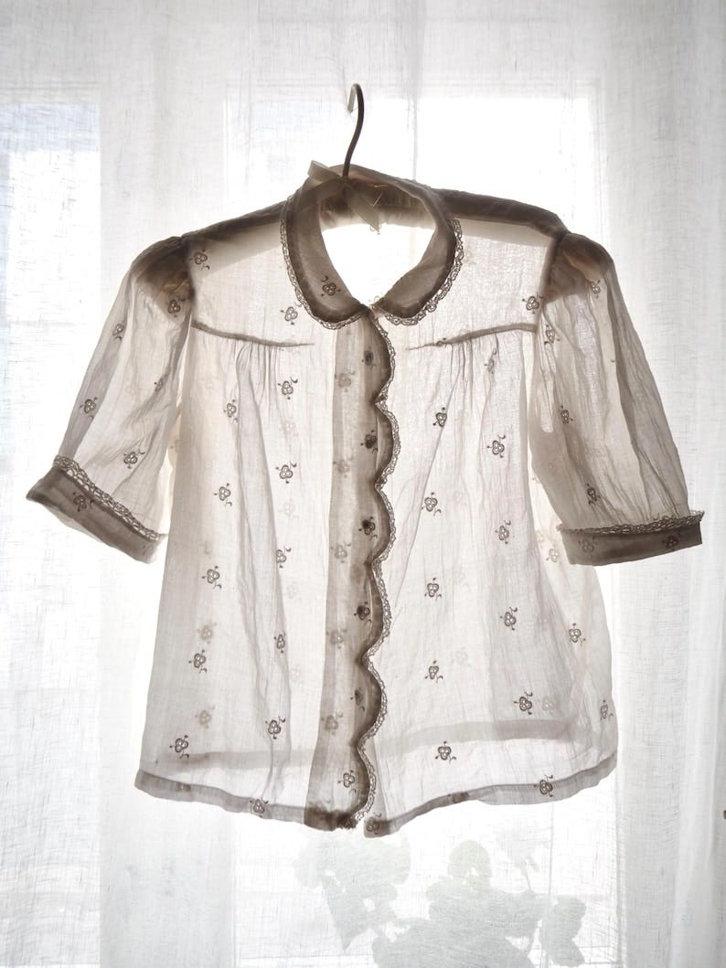 May include: A sheer, off-white blouse with short sleeves, a scalloped edge, and delicate floral embroidery. The blouse has a rounded collar and is buttoned down the front. The sleeves and collar are trimmed with lace. The blouse hangs on a metal hanger.