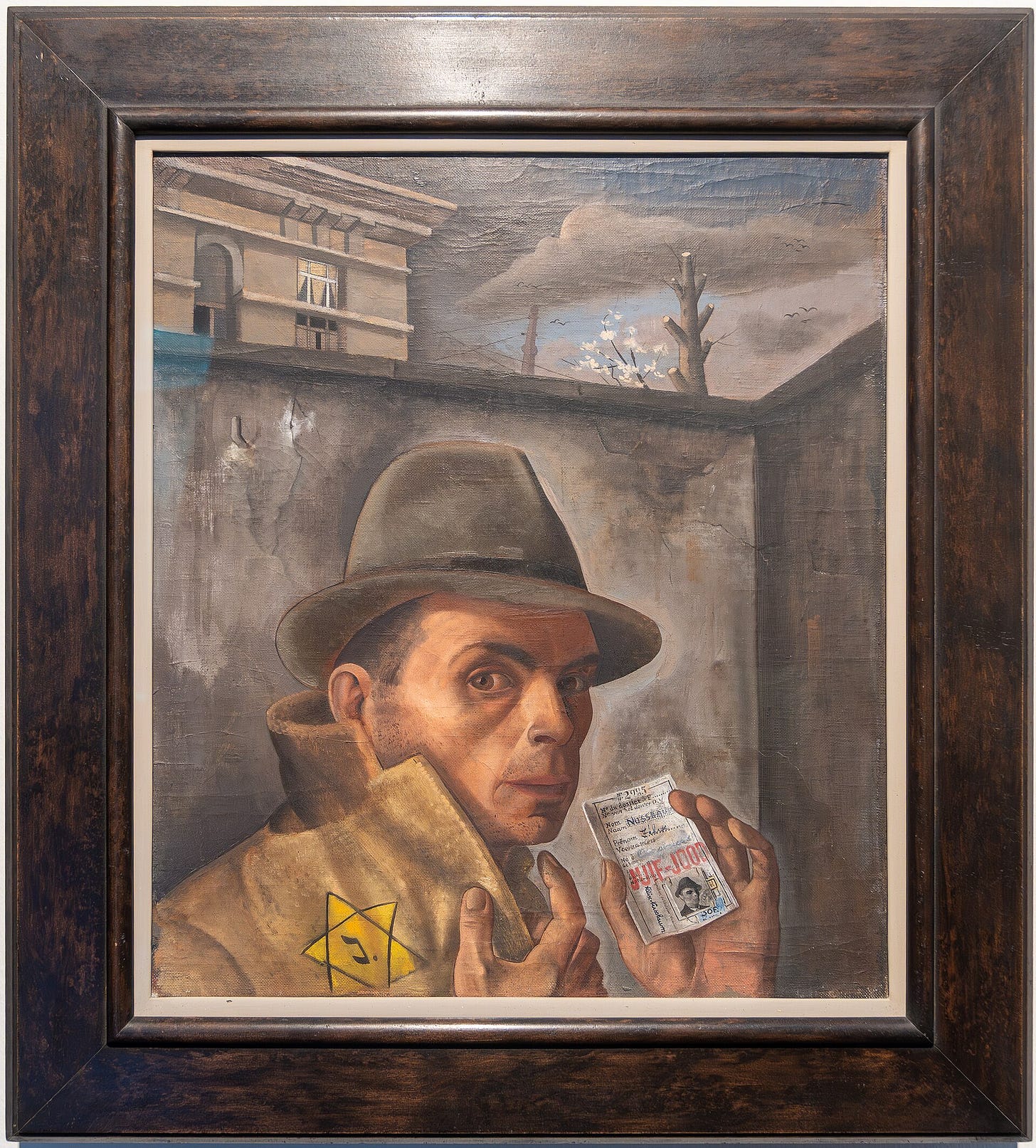 A man in a felt hat turns up his coat collar to show a yellow star marked J and holds up an identity card stamped JUIF-JOOD, in front of a high gray wall with a bare tree and a branch in blossom behind it. A man in a felt hat turns up his coat collar to show a yellow star marked J and holds up an identity card stamped JUIF-JOOD, in front of a high gray wall with a bare tree and a branch in blossom behind it.