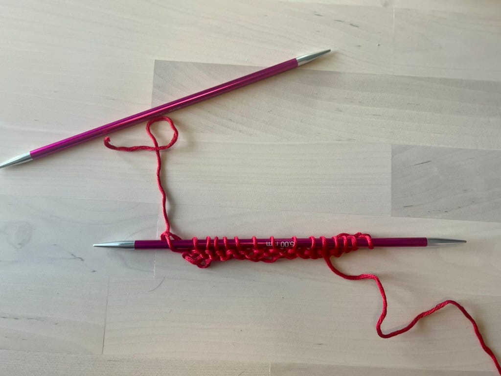 A knitting needle with several rows of red waste yarn knitted, on a wooden surface.