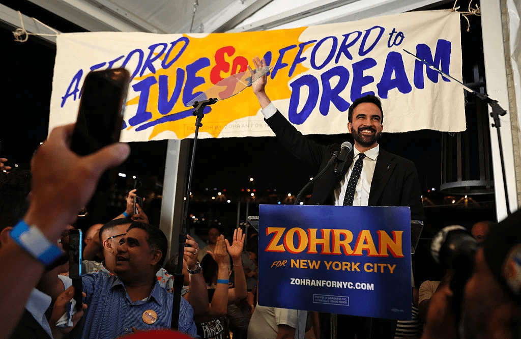 Zohran Mamdani on a podium greeting a crowd of his supporters on election night.