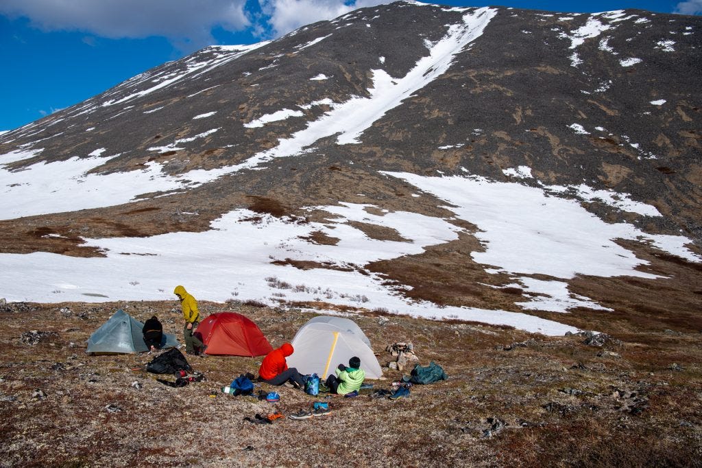Three small tents stand on brown tundra at the foot of a long scree slope streaked with snow, several hikers sitting and moving among scattered gear under a blue sky.