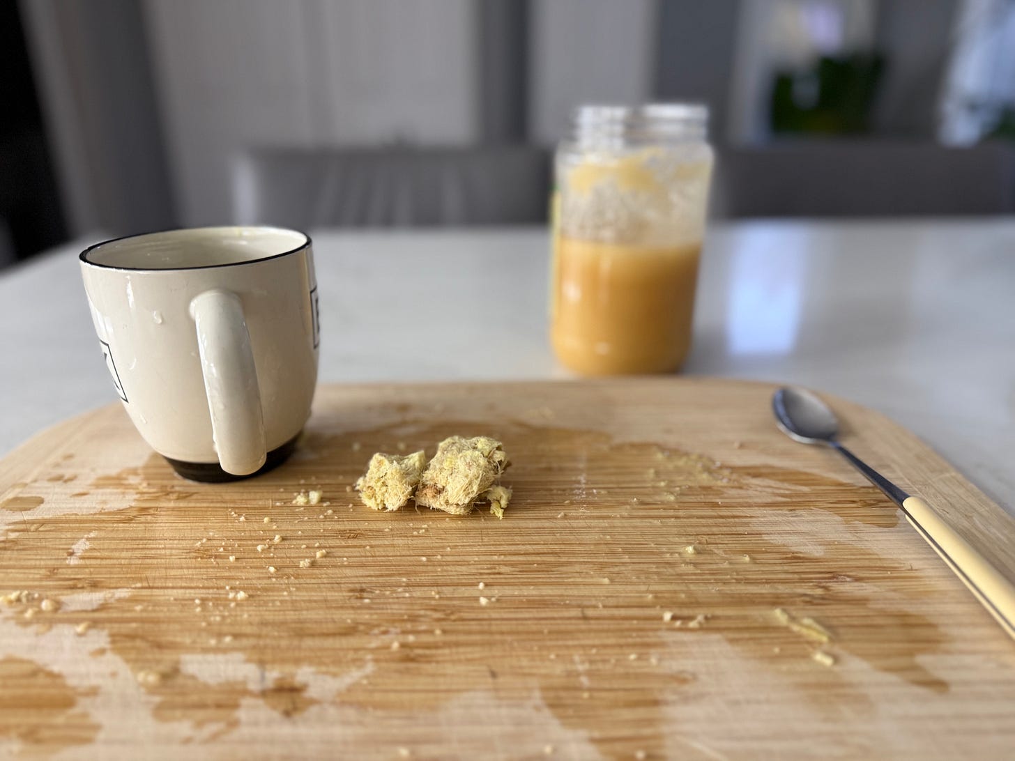 Wooden cutting board with a used mug, scattered ginger pieces, and a jar of golden ginger-honey tea, showing a just-made throat-soothing drink.